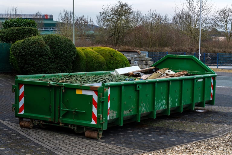 A green dumpster is located in an urban setting, filled with various construction debris and materials.