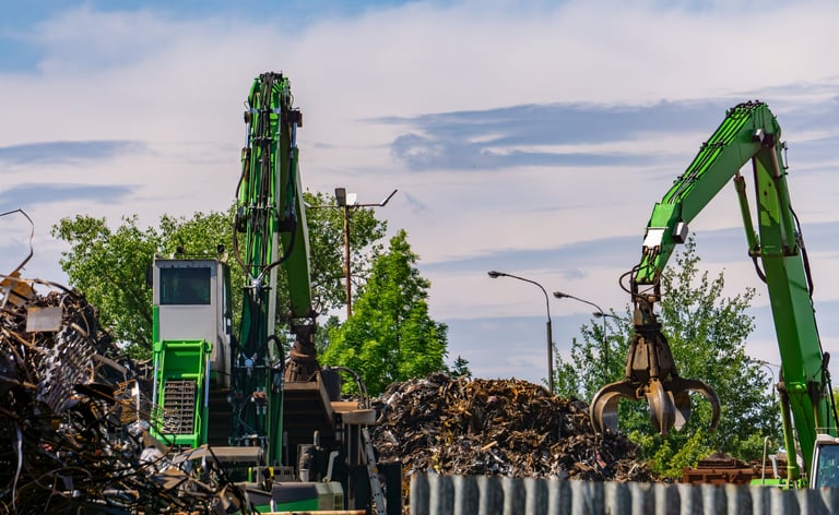 scrapyard featuring two green material handling machines equipped with metal grapples. Piles of scrap metal are visible in the foreground.