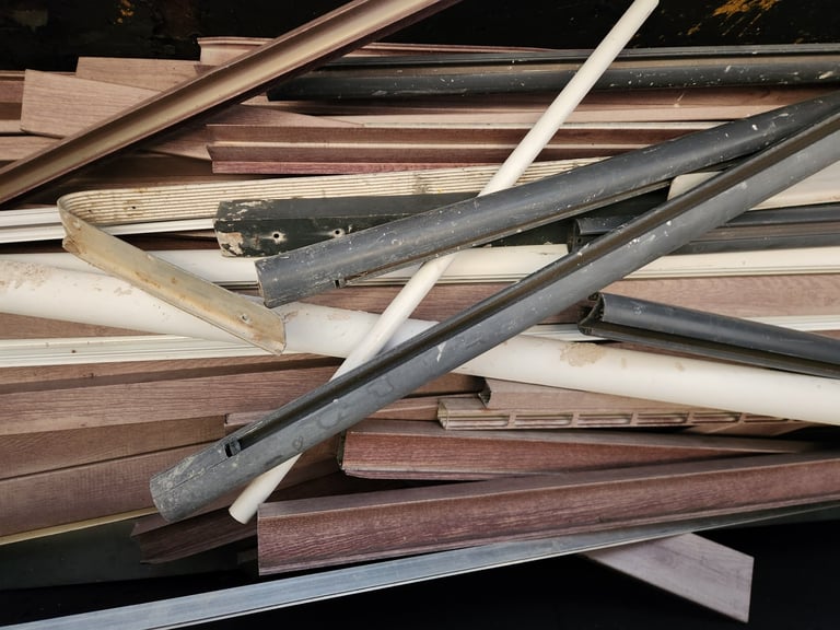 Assorted wooden boards and metal rails stacked together, showing various wood tones and finishes in a lumber or construction pile